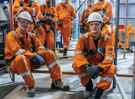 A group portrait of seven construction workers in orange high-visibility coveralls and white hard hats.